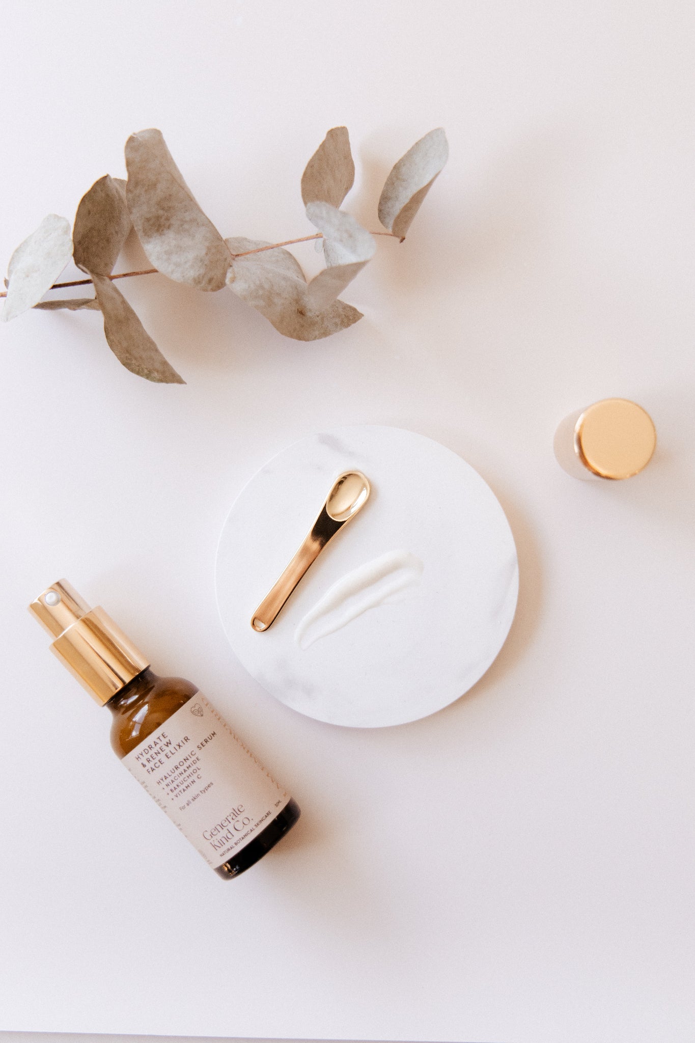 Generate Kind Co. Face Elixir Serum bottle, gold spoon, and eucalyptus leaves on a white surface