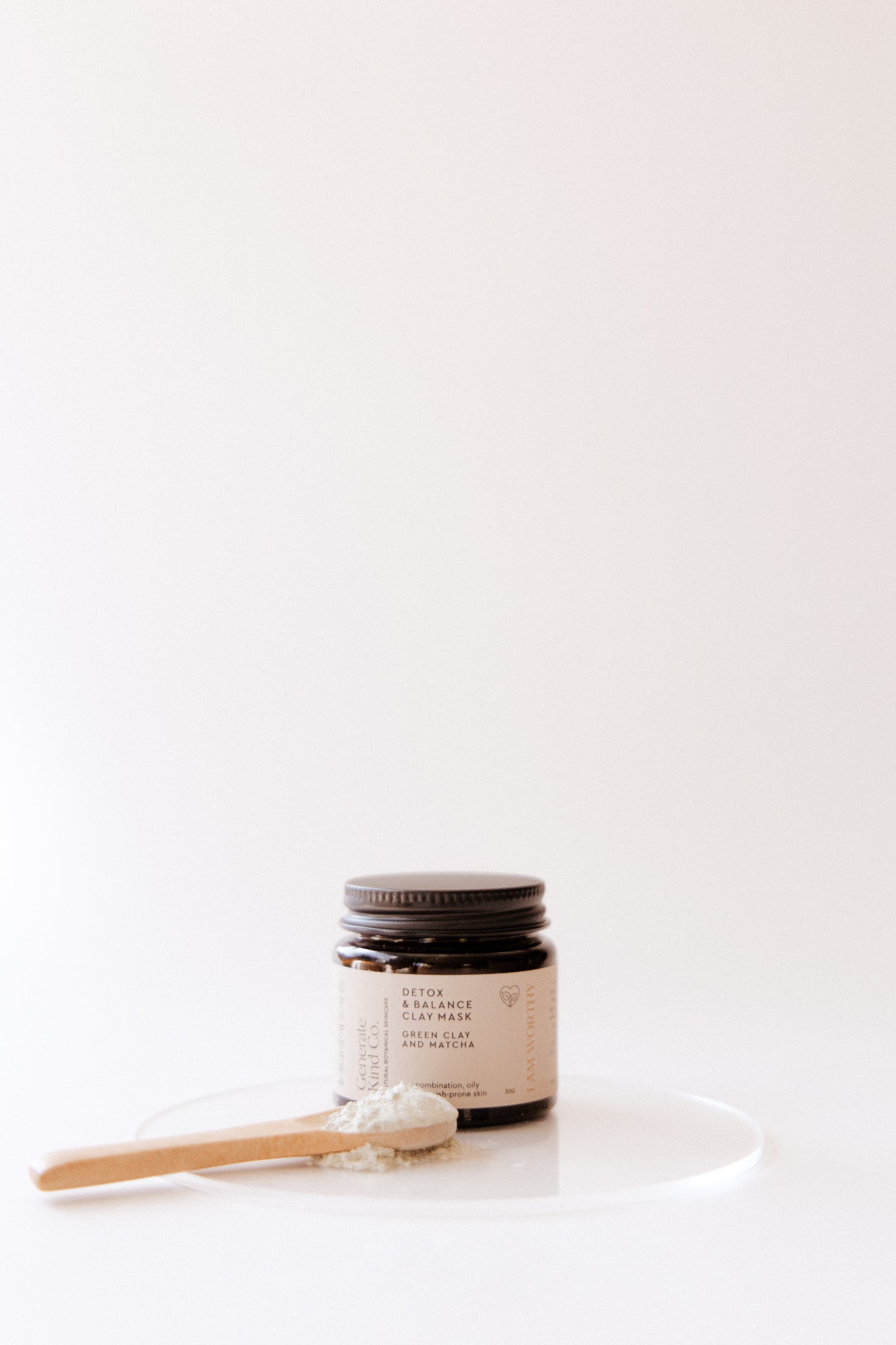 Jar of Green Clay & Match Face Mask with a wooden spoon on a white plate against a white background