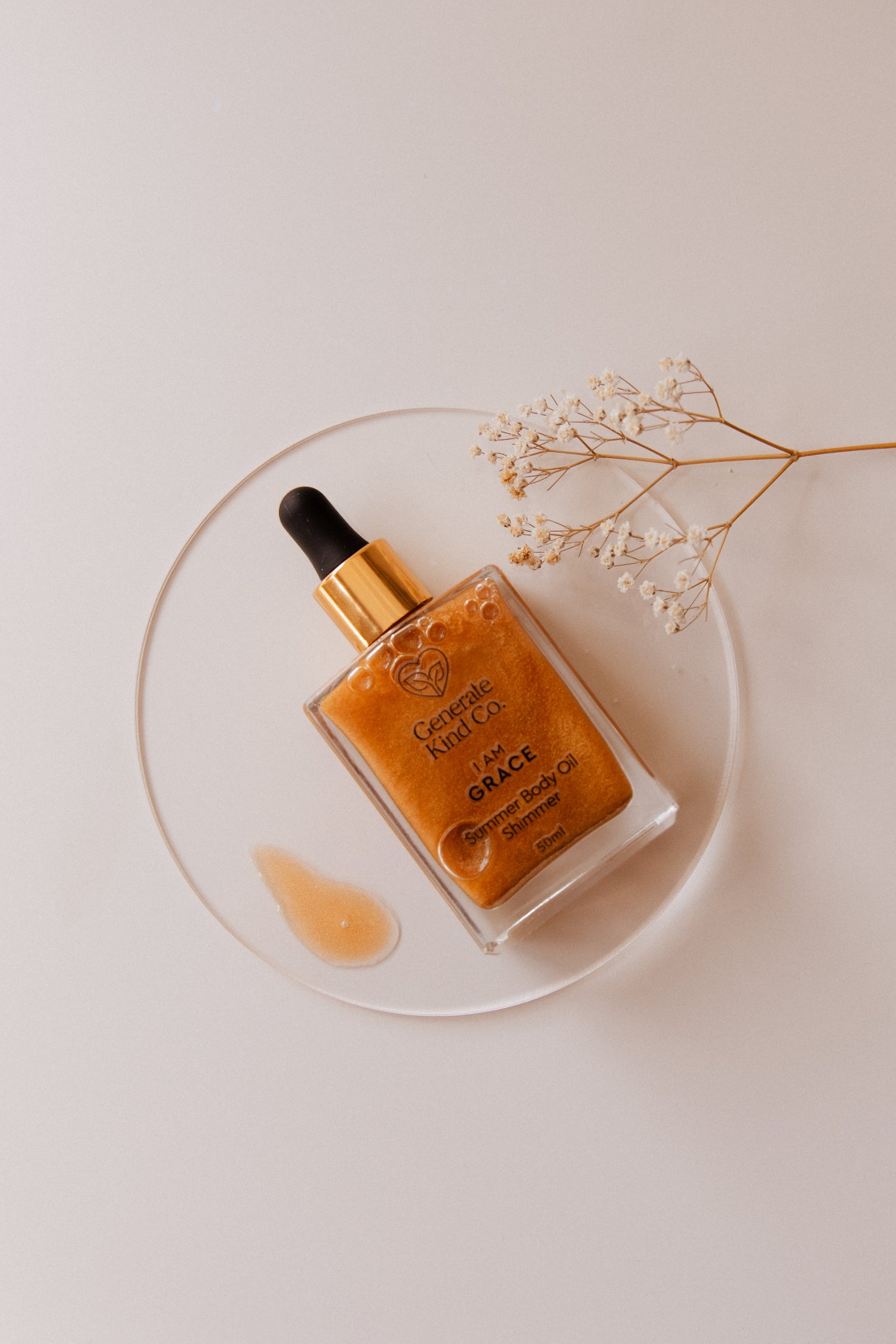 Shimmer Body Oil bottle with dropper on a clear plate with white flowers on a light background
